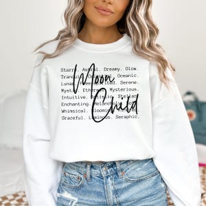 May include: White sweatshirt with black text that reads "Starry. Astral. Dreamy. Glow. Tranquil. Celestial. Oceanic. Moon Child. Lunar. Free-spirited. Serene. Mystic. Ethereal. Mysterious. Intuitive. Artistic. Stellar. Enchanting. Melancholy. Soft. Whimsical. Blooming. Kindred. Graceful. Luminous. Seraphic."