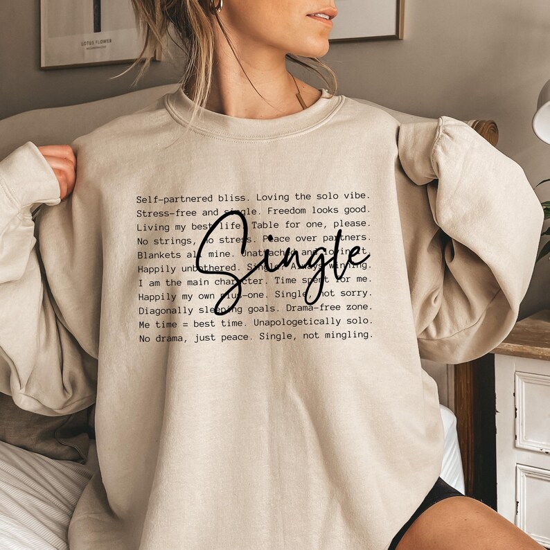 May include: Beige sweatshirt with a black print that reads "Self-partnered bliss. Loving the solo vibe. Stress-free and single. Freedom looks good. Living my best life. Table for one, please. No strings, no stress. Peace over partners. Blankets all mine. Unattached and unbothered. Single. I am the main character. Time spent for me. Happily my own. Single, not sorry. Diagonally sleeping goals. Drama-free zone. Me time = best time. Unapologetically solo. No drama, just peace. Single, not mingling."