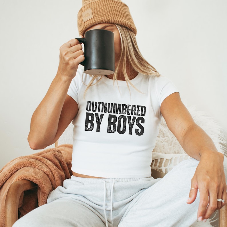 May include: White crop top with the text "OUTNUMBERED BY BOYS" in black. The person in the image is wearing a tan beanie and holding a black mug. They are also wearing gray sweatpants.