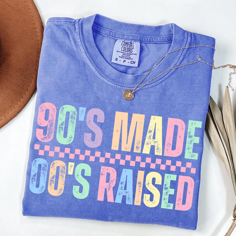 May include: A periwinkle blue t-shirt with the text "90'S MADE 00'S RAISED" in pastel colors. The shirt has a crew neck and short sleeves. A gold necklace with a small pendant rests on the shirt.