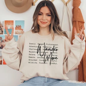 K&ouml;nnte beinhalten: Beiges Sweatshirt mit den Worten "Wander More" in schwarzer Schrift. Das Sweatshirt hat auch eine Liste von W&ouml;rtern wie "Scenic", "Rugged" und "Adventurous". Das Model l&auml;chelt und zeigt das Friedenszeichen.