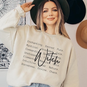 Peut inclure: Un sweat-shirt à manches longues de couleur crème avec un texte noir imprimé : Enchanting. Sacred. Pure. Mystical. Grounded. Calm. Commanding. Spellbinding. Unstoppable. Flourishing. Elemental. Magical. Bold. Intuitive. Lunar. Nature. Sorceress. Divine. Charm. Witch.