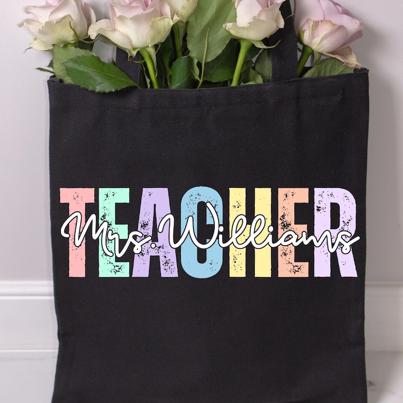 K&ouml;nnte beinhalten: Schwarze Tragetasche mit dem Wort "TEACHER" in bunten, verwitterten Blockbuchstaben, mit "Mrs. Williams" in Schreibschrift. Die Tasche ist mit rosa Rosen gef&uuml;llt.