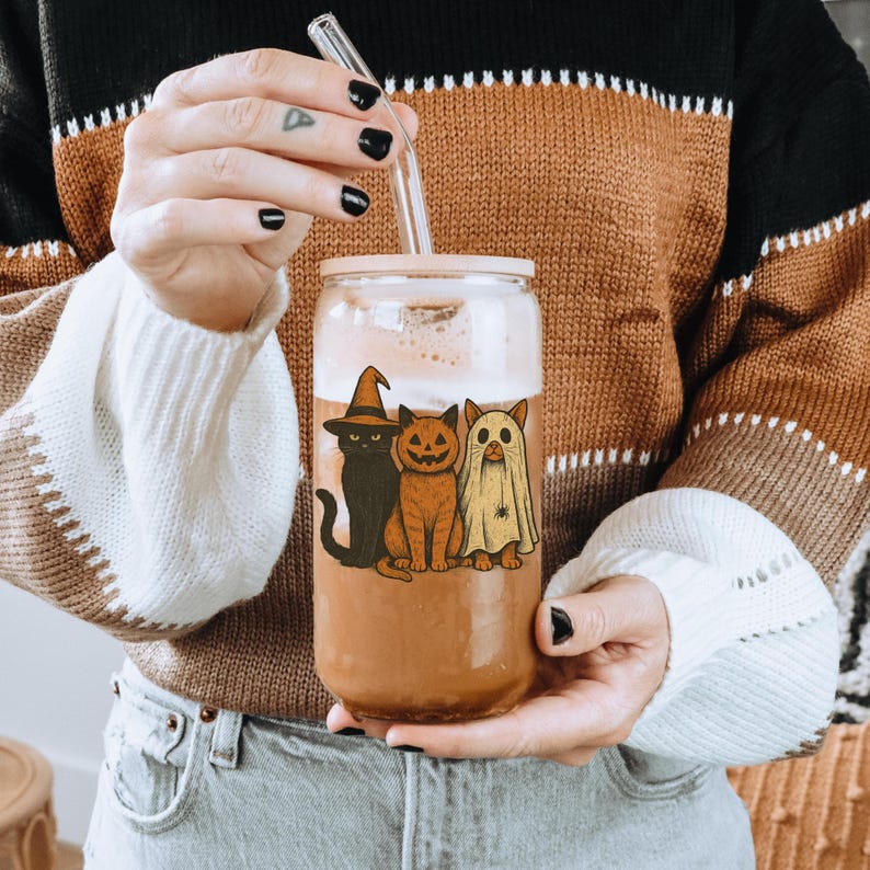 K&ouml;nnte beinhalten: Ein klares Glasgetr&auml;nk mit einem hellrosa Deckel und einem Glasstrohhalm. Der Becher hat ein Halloween-Design mit einer schwarzen Katze mit Hexenhut, einer orangefarbenen Katze mit K&uuml;rbisgesicht und einer Geisterkatze. Der Becher ist mit einem braunen Getr&auml;nk gef&uuml;llt.