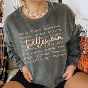 May include: Dark gray sweatshirt with the word "Halloween" in a cursive font. The sweatshirt features a list of Halloween-themed words in a light orange color, including "Spooky," "Creepy," and "Mysterious."