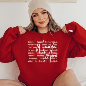 K&ouml;nnte beinhalten: Ein rotes Sweatshirt mit dem wei&szlig;en Schriftzug "Wander More". Das Sweatshirt hat auch eine Liste von W&ouml;rtern wie "Scenic", "Adventurous" und "Majestic". Die Person auf dem Bild tr&auml;gt eine beige M&uuml;tze.