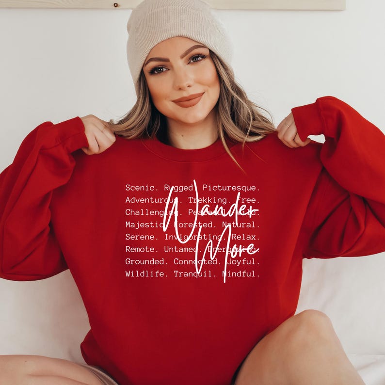 K&ouml;nnte beinhalten: Ein rotes Sweatshirt mit dem wei&szlig;en Schriftzug "Wander More". Das Sweatshirt hat auch eine Liste von W&ouml;rtern wie "Scenic", "Adventurous" und "Majestic". Die Person auf dem Bild tr&auml;gt eine beige M&uuml;tze.