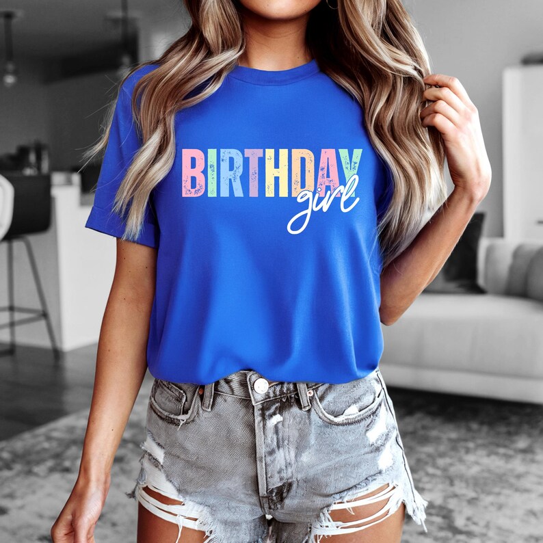 Może przedstawiać: Kr&oacute;lewsko niebieska koszulka z napisem "BIRTHDAY girl" wydrukowanym z przodu w kolorowych pastelowych literach i czcionce. Koszulka jest z kr&oacute;tkim rękawem i wydaje się być wykonana z miękkiego materiału.
