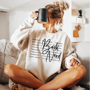 May include: A white sweatshirt with black text that reads "Bookworm. Page Turner. Informed. Word Lover. Novelist. Book Buff. Imaginative. Thoughts. Novel Love. Literary Enthusiast. Book Wisdom. Paperback Pro. She. Collection. Chapter Chase. Fiction Fan. Readaholic. Story Seeker. Smart. Book Hunter. Bright. Reflecting."