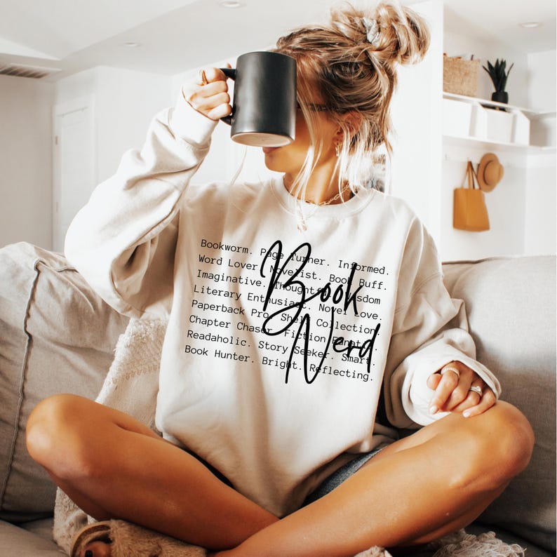 May include: A white sweatshirt with black text that reads "Bookworm. Page Turner. Informed. Word Lover. Novelist. Book Buff. Imaginative. Thoughts. Novel Love. Literary Enthusiast. Book Wisdom. Paperback Pro. She. Collection. Chapter Chase. Fiction Fan. Readaholic. Story Seeker. Smart. Book Hunter. Bright. Reflecting."