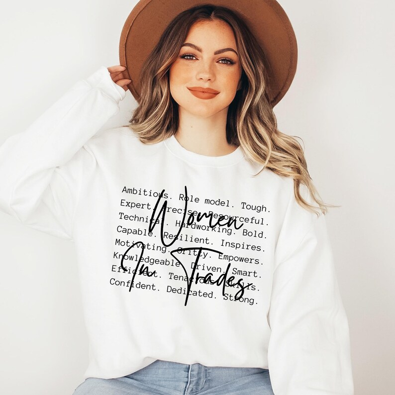 May include: White sweatshirt with black text that reads "Ambitious. Role model. Tough. Expert. Resourceful. Technical. Hardworking. Bold. Capable. Resilient. Inspires. Motivating. Gritty. Empowers. Knowledgeable. Driven. Smart. Efficient. Tenacious. Confident. Dedicated. Strong. Women in Trades"