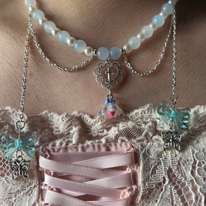 May include: A delicate layered necklace with a white pearl and blue glass bead chain. The necklace features a silver heart pendant with a cross, a small pink and white charm, and two blue butterfly charms.