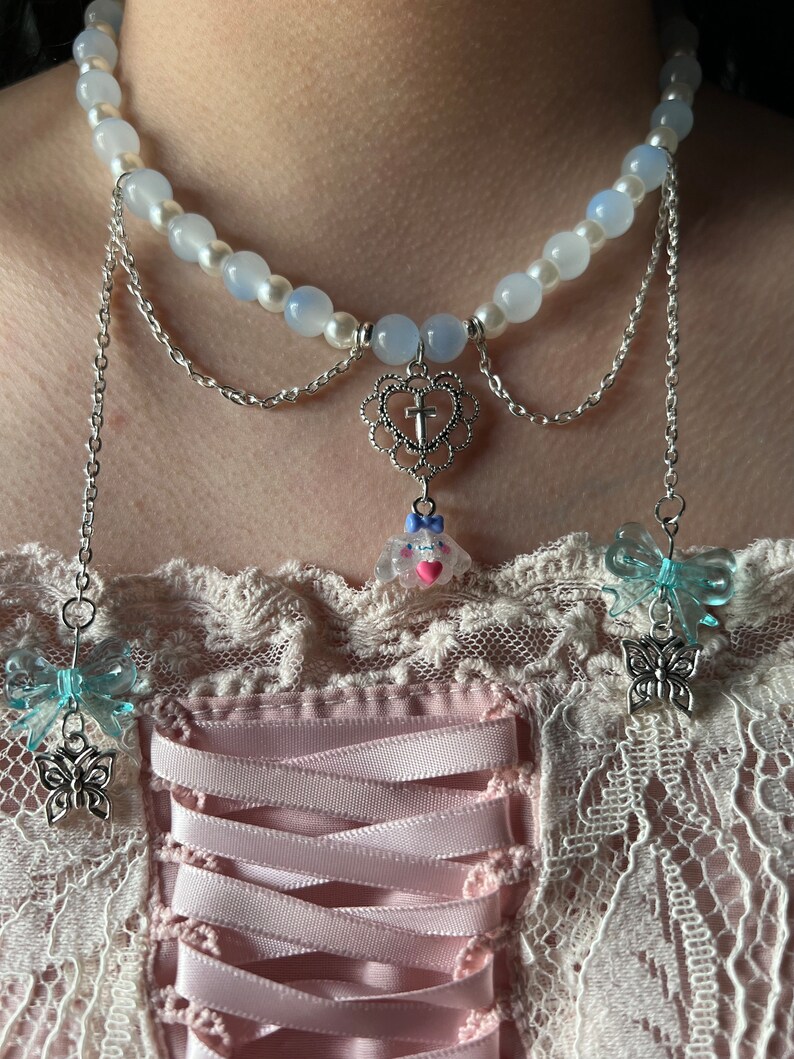 May include: A delicate layered necklace with a white pearl and blue glass bead chain. The necklace features a silver heart pendant with a cross, a small pink and white charm, and two blue butterfly charms.