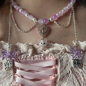May include: A layered necklace with a delicate silver chain and a purple and white beaded choker. The choker features a heart charm with a cross and a small unicorn charm. The necklace has two dangling butterfly charms with purple bows.