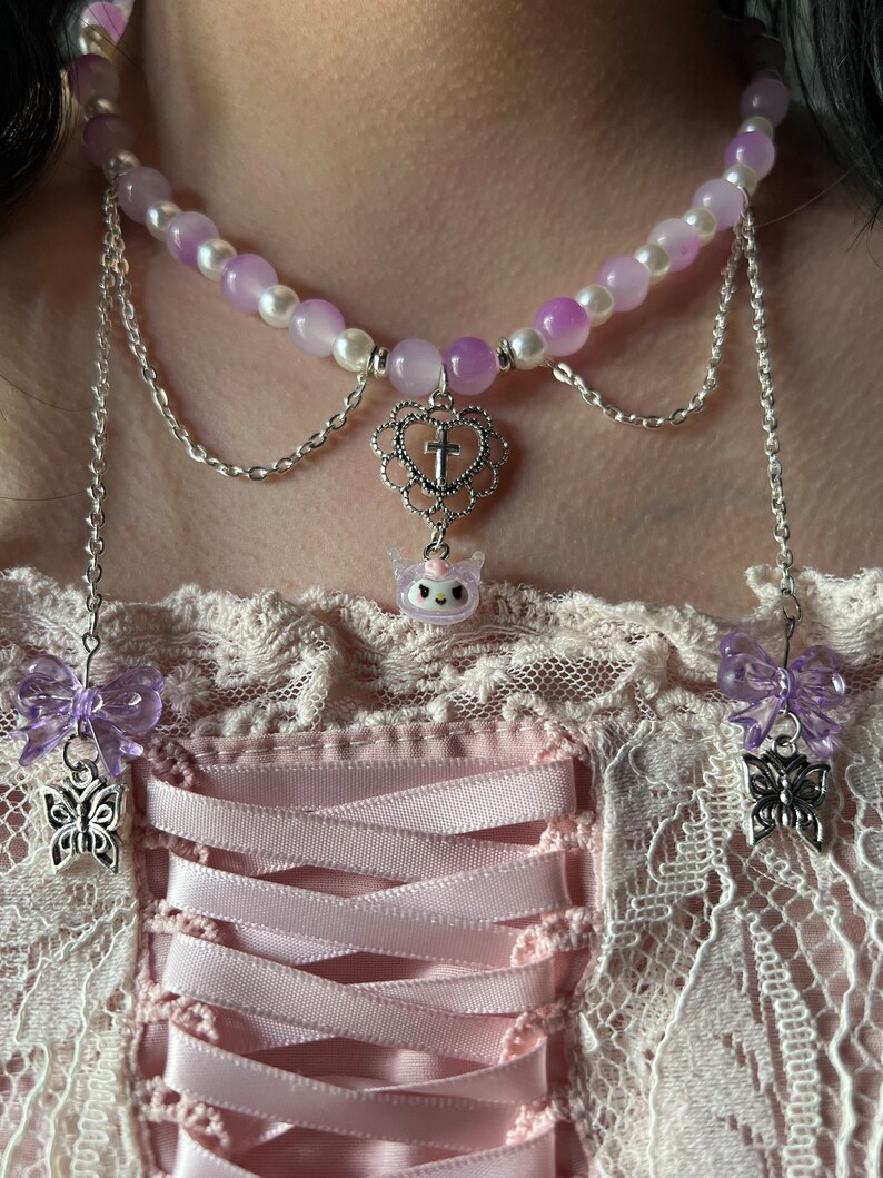 May include: A layered necklace with a delicate silver chain and a purple and white beaded choker. The choker features a heart charm with a cross and a small unicorn charm. The necklace has two dangling butterfly charms with purple bows.