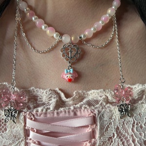 May include: A pink and white beaded choker necklace with a silver chain and a charm featuring a cartoon character with a cross. The necklace has two dangling silver chains with pink butterfly charms.