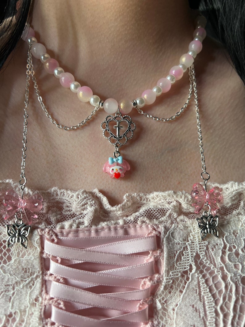 May include: A pink and white beaded choker necklace with a silver chain and a charm featuring a cartoon character with a cross. The necklace has two dangling silver chains with pink butterfly charms.