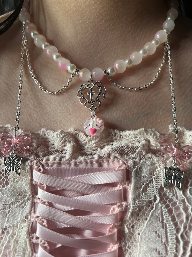 May include: A layered necklace with a pink and white pearl strand, silver chains, and a charm with a cross in a heart and a Hello Kitty charm. The necklace is worn over a pink and white lace top with a pink ribbon.
