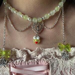 May include: A layered silver chain necklace with a green and white beaded choker, a heart charm with a cross, and a green frog charm. The necklace is worn over a pink and white lace top.