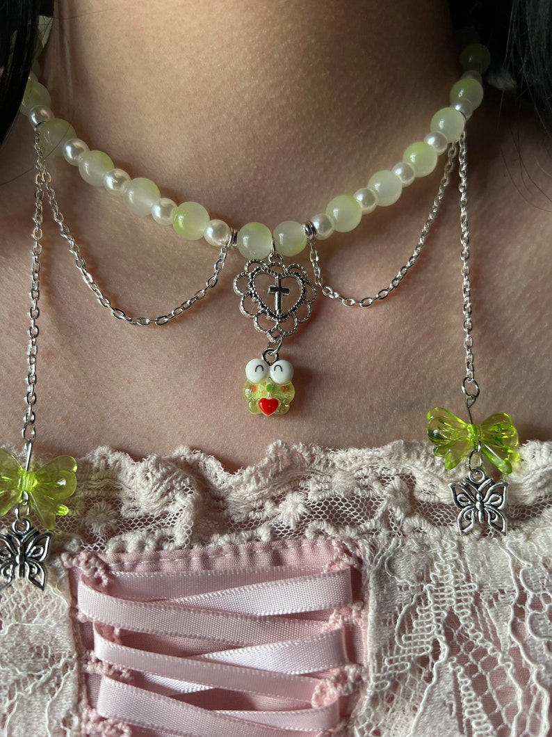 May include: A layered silver chain necklace with a green and white beaded choker, a heart charm with a cross, and a green frog charm. The necklace is worn over a pink and white lace top.