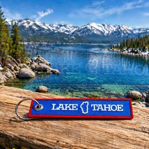 Puede incluir: Un llavero rectangular de tela azul con borde rojo y una anilla de metal plateado. El texto LAKE TAHOE está bordado en blanco, con un pequeño contorno blanco de la forma del lago entre las dos palabras en la parte frontal.