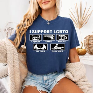 Pode incluir: Camiseta azul marinho com o texto "I SUPPORT LGBTQ" e ícones gráficos representando licor, armas, cerveja, seios e rapidinhas. A camiseta é feita de um material macio.