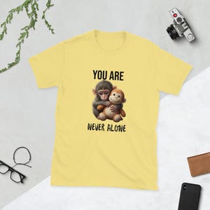 May include: A pale yellow t-shirt with a graphic of two hugging monkeys and the text "YOU ARE NEVER ALONE" in black. The monkeys are illustrated in a cartoon style with detailed shading.