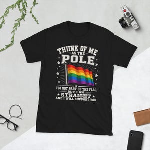 Puede incluir: Una camiseta negra de manga corta con un gráfico de una bandera arcoíris en un mástil. El texto dice: THIINK OF ME AS THE POLE I'M NOT PART OF THE FLAG, BUT I AM STRAIGHT AND I WILL SUPPORT YOU. El diseño incluye estrellas blancas y líneas radiantes.