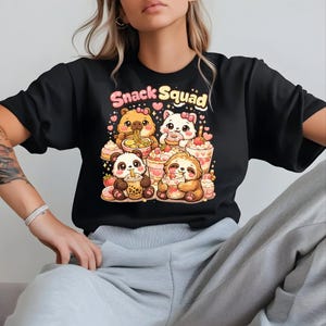 Puede incluir: Camiseta negra con un gráfico de dibujos animados de un oso, un gato, un panda y un perezoso disfrutando de aperitivos. El texto "Snack Squad" está encima de los personajes. El diseño incluye corazones, donuts y bebidas.