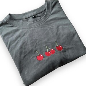 May include: A folded, dark gray t-shirt featuring an embroidered design of three red cherries with green stems. The cherries are positioned horizontally across the front of the shirt. The shirt is a simple crew neck style.