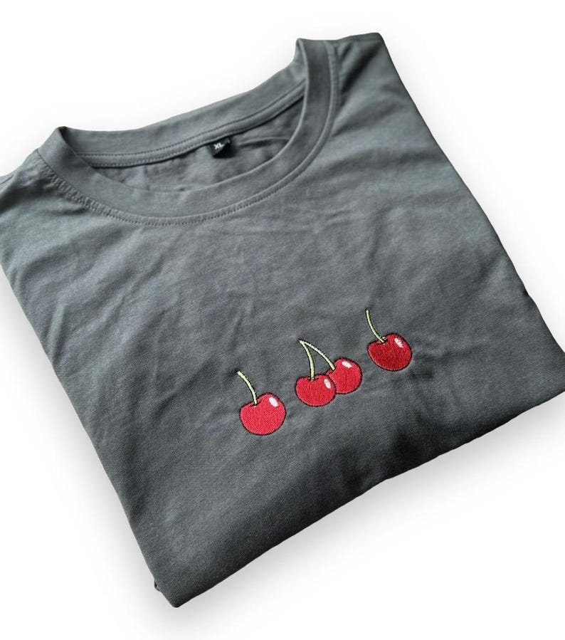 May include: A folded, dark gray t-shirt featuring an embroidered design of three red cherries with green stems. The cherries are positioned horizontally across the front of the shirt. The shirt is a simple crew neck style.
