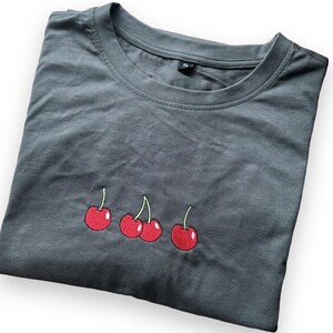 May include: A folded charcoal gray t-shirt with a crew neck. The shirt features an embroidered design of three red cherries with green stems. The shirt is made of a soft material and is a size XL.