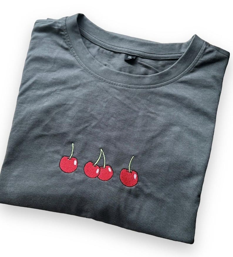 May include: A folded charcoal gray t-shirt with a crew neck. The shirt features an embroidered design of three red cherries with green stems. The shirt is made of a soft material and is a size XL.