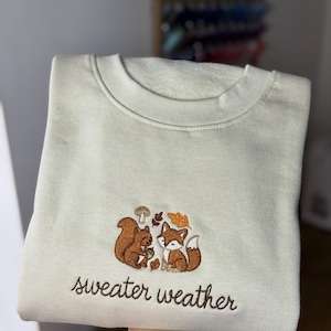 May include: Cream-coloured sweatshirt with embroidered design featuring a squirrel and fox, accompanied by autumn leaves and a mushroom. The phrase "sweater weather" is embroidered below the design in brown thread.
