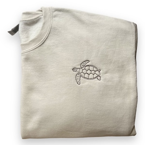 Könnte beinhalten: Ein gefaltetes, hellbeige Sweatshirt mit Rundhalsausschnitt. Auf der Vorderseite des Sweatshirts befindet sich eine detaillierte Stickerei einer Meeresschildkröte in braunem Garn. Das Kleidungsstück ist aus weichem Material gefertigt.