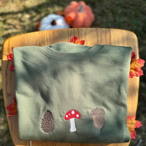 May include: An olive green sweatshirt featuring embroidered autumnal motifs. The designs include a pinecone, a mushroom with a red cap and white spots, and an acorn. The sweatshirt is folded on a wooden surface, with autumn leaves.