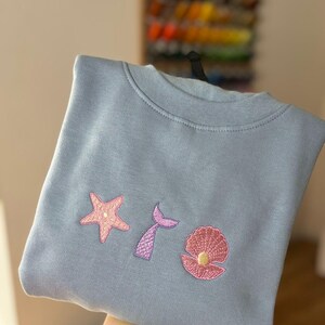 May include: A light blue sweatshirt featuring embroidered designs of a pink starfish, a purple mermaid tail, and a pink seashell with a yellow pearl. The sweatshirt is displayed on a black hanger.