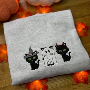 May include: A light grey sweatshirt with embroidered Halloween designs. The designs include a black cat wearing a witch's hat, a white ghost, and a black cat with devil horns. The cats have green eyes. The sweatshirt is surrounded by orange autumn leaves.