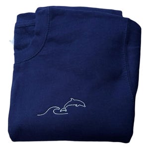 Puede incluir: Sudadera azul marino con un diseño bordado en blanco de un delfín saltando sobre una ola. La sudadera está doblada, mostrando el cuello redondo y el gráfico del delfín en la parte delantera. La tela parece suave y cómoda.