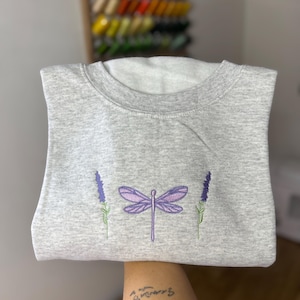 Può includere: Felpa grigia girocollo con un disegno ricamato. Il disegno include una libellula viola e due rametti di lavanda. La felpa è piegata.