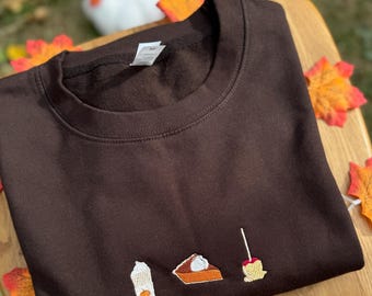 Può includere: Una felpa marrone con ricami a tema autunnale. Il design include un latte speziato alla zucca, una fetta di torta con panna montata e una mela caramellata. La felpa è esposta su una superficie di legno con foglie autunnali.