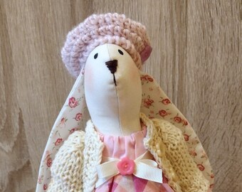 Könnte beinhalten: Eine weiße Hasenpuppe, die ein rosa-weißes Karo-Kleid, eine cremefarbene Strickjacke und eine rosa Strickmütze trägt. Die Puppe hat einen rosa Knopf auf der Brust.