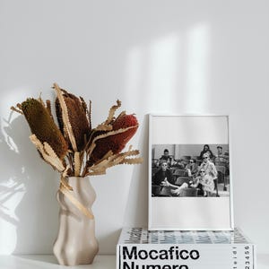 May include: A white vase with a textured surface holds a bouquet of dried brown and red banksia flowers. A black and white photo of a group of people sitting in a row is framed in a white frame and sits on top of a white book with the title "Mocacifico Numero" printed in black.