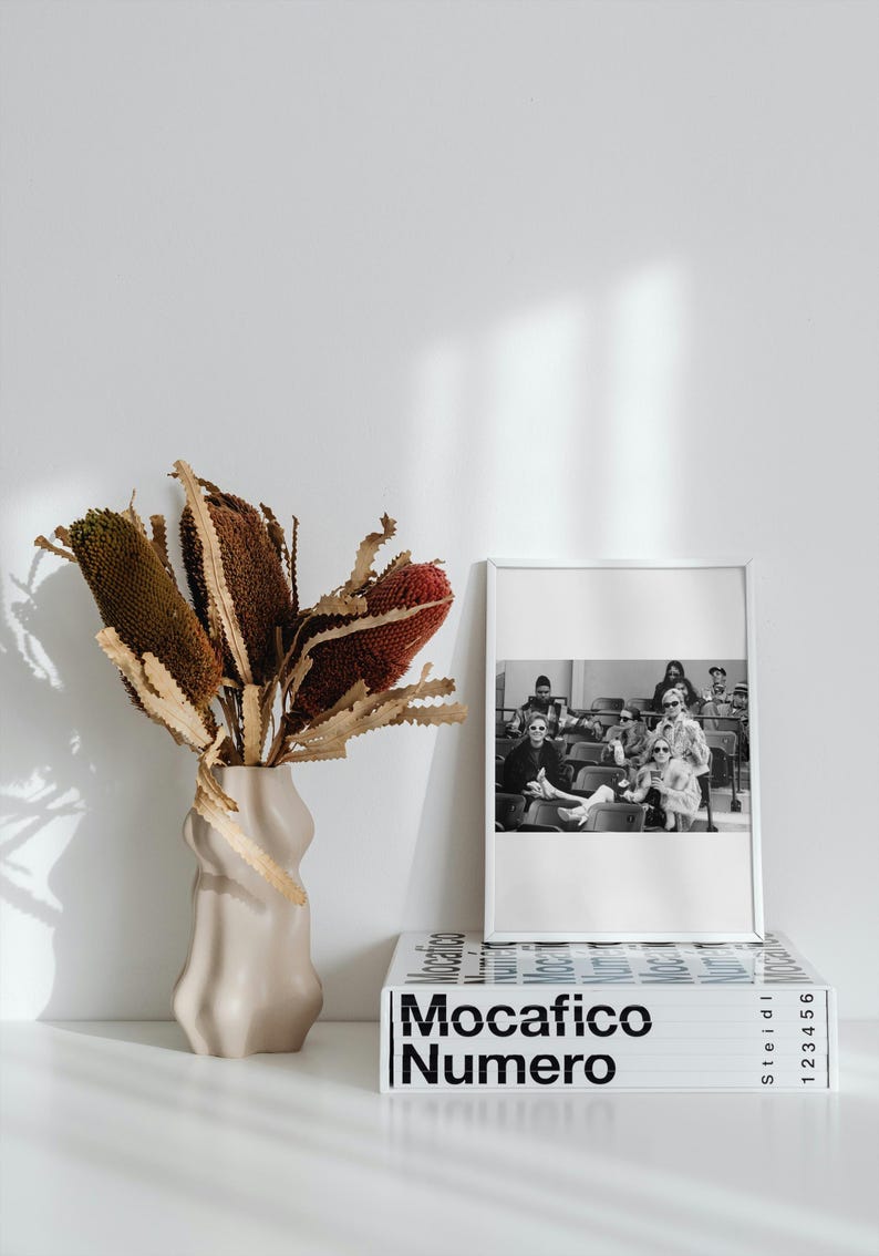 May include: A white vase with a textured surface holds a bouquet of dried brown and red banksia flowers. A black and white photo of a group of people sitting in a row is framed in a white frame and sits on top of a white book with the title "Mocacifico Numero" printed in black.