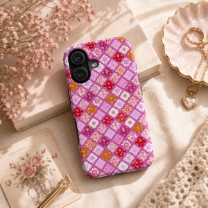 May include: A smartphone case featuring a repeating diamond-shaped patchwork pattern in shades of pink, magenta, red, and orange. The design consists of various floral and geometric motifs within each square, with a black camera lens cutout.