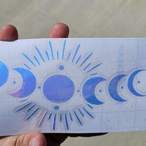 Pode incluir: Um autocolante holográfico com um design celestial com fases da lua e um sol radiante. O autocolante tem um esquema de cores azul e iridescente, com um fundo transparente. O design está num suporte branco com padrão de grelha.