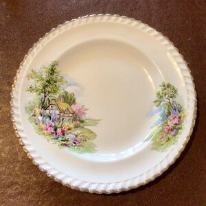 May include: A white ceramic plate with a gold rim features a cottage scene with a green roof, pink flowers, and trees. The plate has a white background with a subtle floral pattern.