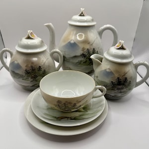 May include: A vintage porcelain tea set comprising a teapot, creamer, sugar bowl, teacup, and saucers. The set is painted with a landscape design of a mountain, trees, and a lake in muted tones. The set has a white base with gold trim.