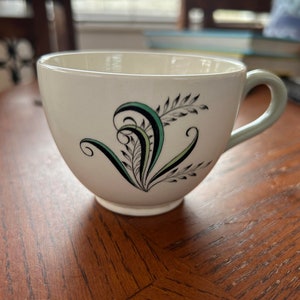 Può includere: Una tazza da tè bianca con un disegno floreale verde e nero. La tazza ha una forma arrotondata e un manico.
