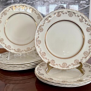 May include: A set of cream-coloured plates with gold rims and decorative gold floral patterns. The plates are stacked on clear stands and a dark wooden surface. The plates have a scalloped edge.